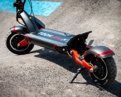 rear diagonal view of a black Techlife X7 with red details leaning on its stand in sand