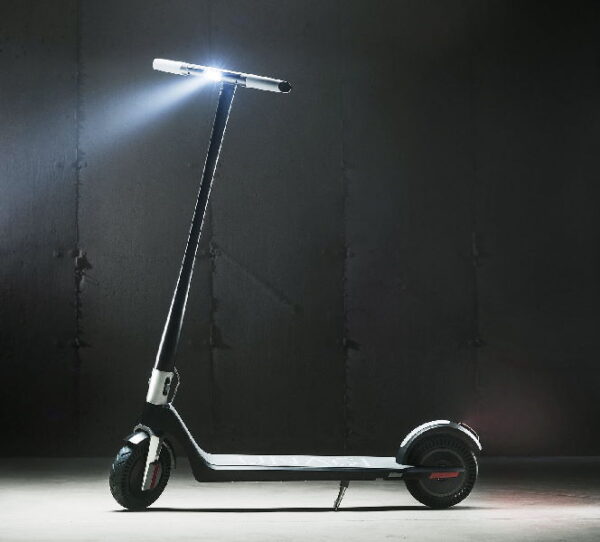 side view of a Unagi Model One electric scooter leaning on its stand with its headlight turned on in a dark room