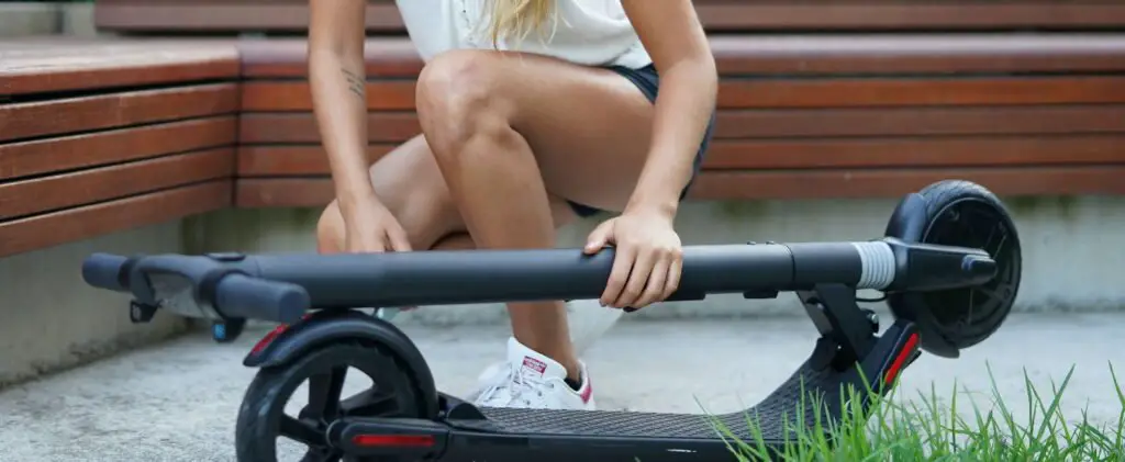 person kneeling over a folded black electric scooter with red details in a park